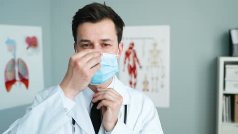 Caucasian-young-male-doctor-talking-to-camera-and-teaching-to-wear-medical-mask-in-cabinet.-Medic-blogger-taking-on-protection-from-coronavirus.-Videoblog-on-healthcare.-Physician-consultation.